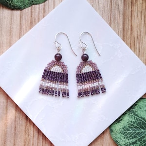 May include: A pair of handmade beaded earrings. Each earring features a dark purple bead at the top, a beaded section in shades of purple, white, and lavender, and a fringe of purple and white beads. The earrings hang from silver-colored hooks. The text "Iris Elm Jewelry" is visible.