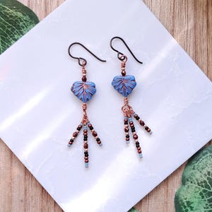 May include: Dangle earrings featuring blue leaf-shaped beads with dark wire-wrapped accents. Each earring includes three dangling strands of small metallic copper, black, and light blue seed beads suspended from a dark metal hook wire.