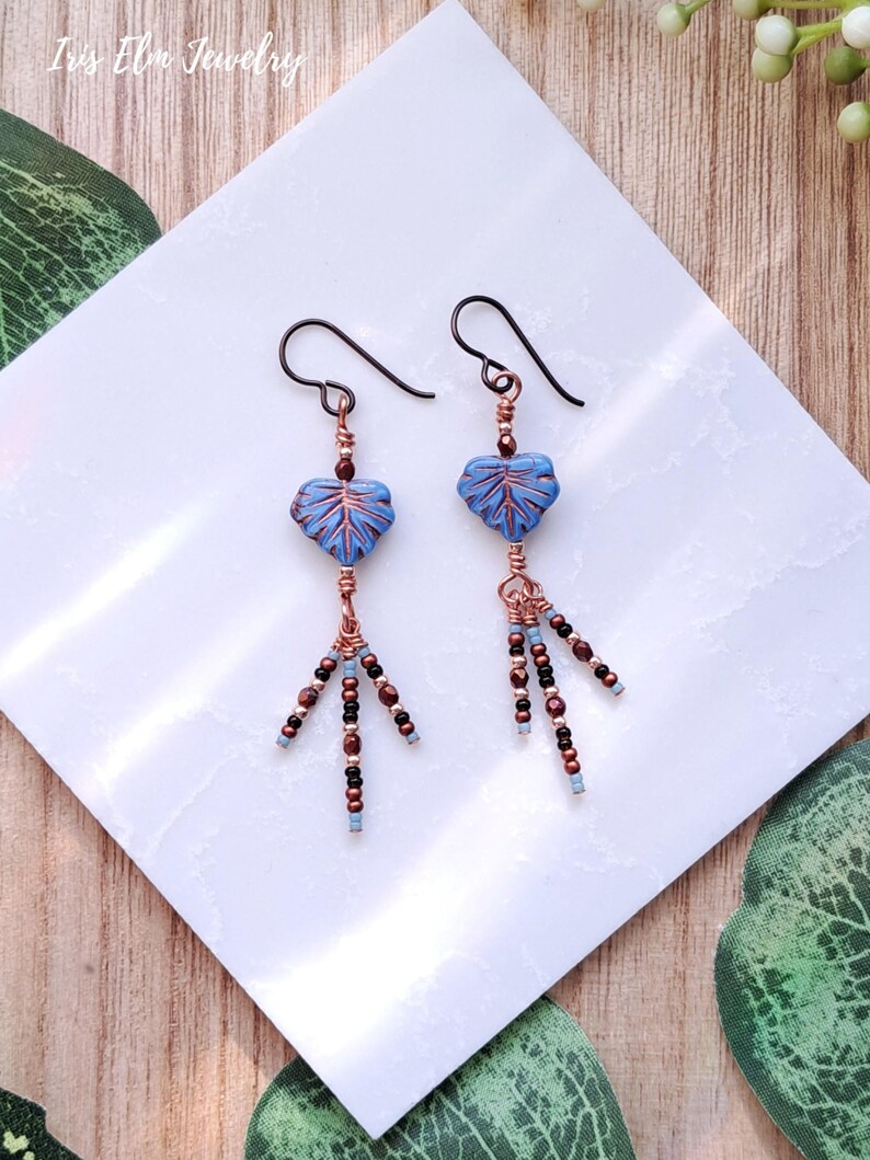 May include: Dangle earrings featuring blue leaf-shaped beads with dark wire-wrapped accents. Each earring includes three dangling strands of small metallic copper, black, and light blue seed beads suspended from a dark metal hook wire.