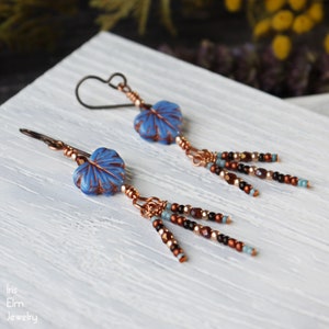 May include: A pair of dangle earrings featuring blue leaf-shaped glass beads with copper-colored veins, suspended from dark metal ear wires. Each earring includes three dangling strands of small copper, black, and light blue glass seed beads.