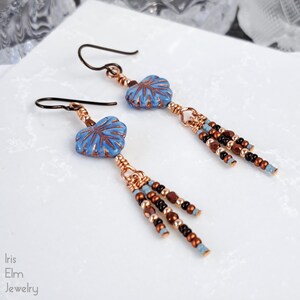 May include: A pair of dangle earrings featuring light blue, leaf-shaped ceramic beads with copper-colored veining. Each earring includes copper wire wrapping and a fringe of small, multi-colored glass beads in shades of copper, black, and blue.