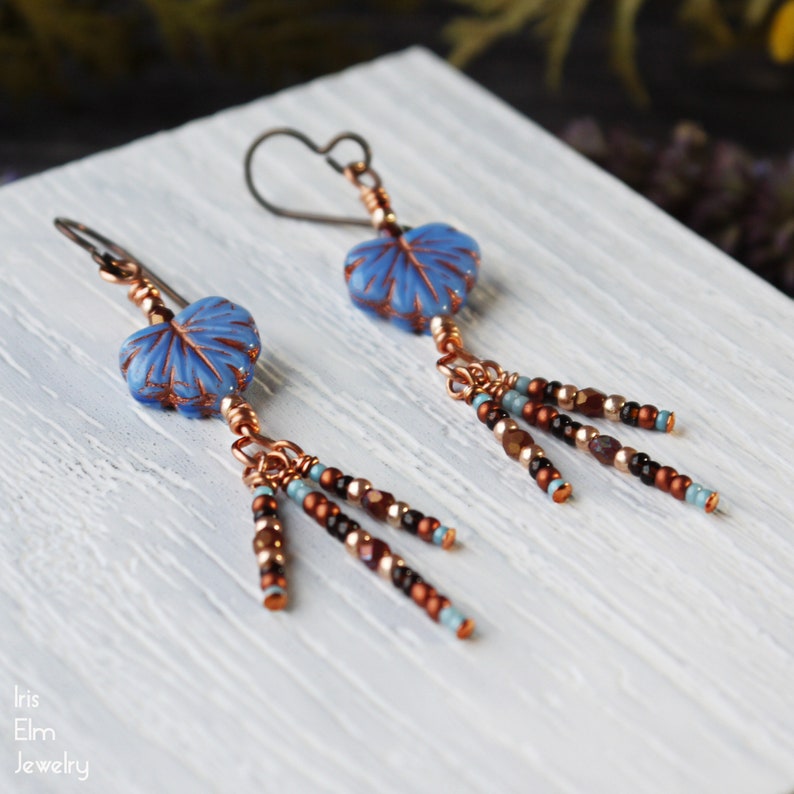 May include: A pair of dangle earrings featuring light blue leaf-shaped glass beads with copper-colored veining. Each earring has a dark metal hook and three dangling strands of small metallic copper, black, and light blue seed beads.
