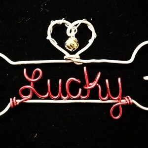May include: A white wire dog bone with a red wire name "Lucky" and a gold bell hanging from a white wire heart.