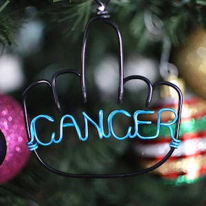 May include: A black wire ornament shaped like a middle finger with the word "CANCER" spelled out in blue wire. The ornament is hanging on a Christmas tree with other ornaments.