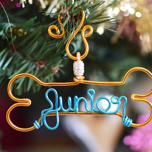 May include: A gold-colored wire dog bone ornament with the name "Junior" in blue wire. The ornament is hanging on a Christmas tree. The ornament has a decorative bead and loop at the top.