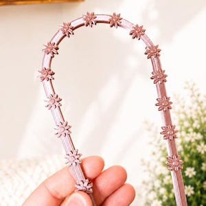 Puede incluir: Una diadema de color rosa palo con una serie de pequeñas flores tridimensionales espaciadas uniformemente a lo largo del borde exterior. La banda tiene una superficie texturizada y acanalada con una estructura curva y flexible.