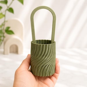 Può includere: Un contenitore cilindrico color verde oliva caratterizzato da una superficie esterna con una texture a coste verticali ondulate. L'oggetto presenta un'alta impugnatura semicircolare ad arco che si estende verso l'alto dal bordo superiore.