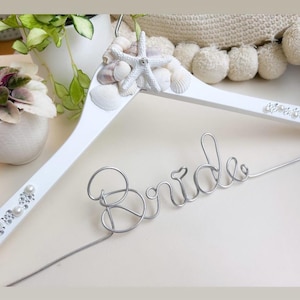 May include: White wooden hanger with a silver wire "Bride" inscription, decorated with seashells and faux pearls.