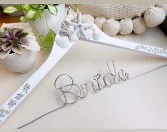 May include: White wooden hanger with a silver wire "Bride" inscription, decorated with seashells and faux pearls.