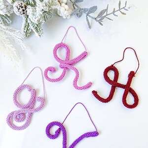 May include: Four knitted ornaments in the shape of letters. The letters are "G", "R", "H", and "V". The ornaments are pink, purple, and red.
