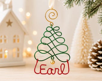 May include: A Christmas tree ornament crafted from green wire in a swirling pattern, topped with a gold wire spiral and a cream ribbon. The base is formed by red wire spelling the name Earl, accented with a small gold bead at the centre.