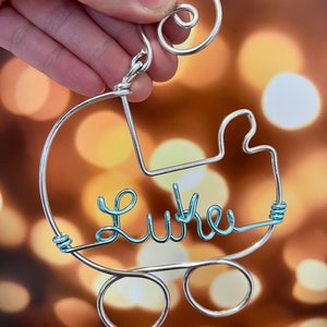 May include: A silver wire ornament shaped like a baby carriage with the name "Luke" written in light blue wire.