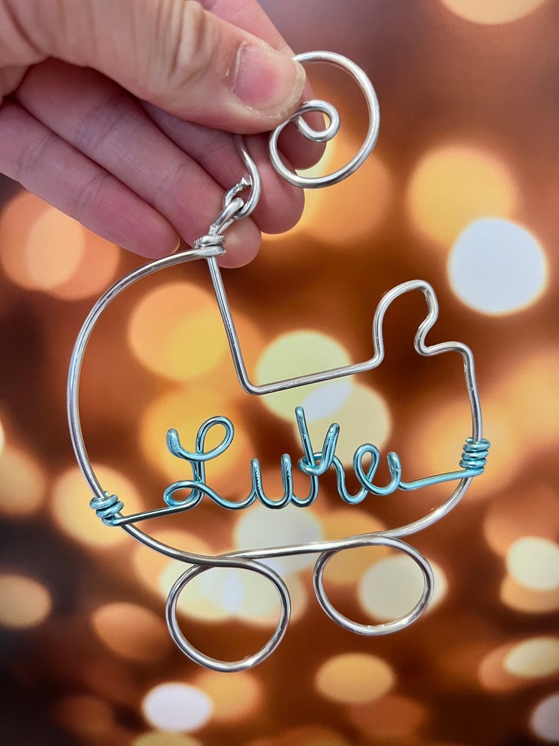 May include: A silver wire ornament shaped like a baby carriage with the name "Luke" written in light blue wire.