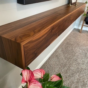 May include: A dark brown wooden floating shelf with a drawer. The shelf is mounted on a white wall and has a natural wood grain finish.
