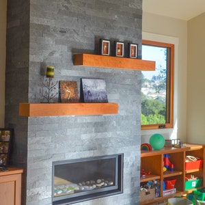 May include: A fireplace with a gray stone surround and a wood mantel. The fireplace has a black metal frame and a glass front. There are two shelves above the fireplace, one with a clock and a small metal tree with candles, and the other with three small framed pictures. The text "Shown In: Classic Cherry" is visible in the upper right corner of the image.