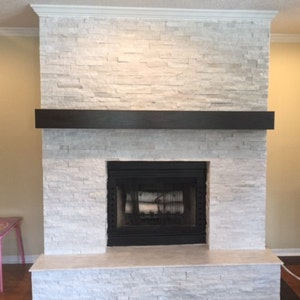 May include: A white stone fireplace with a black mantel and a black fireplace insert. The fireplace is surrounded by white stone tiles.