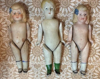 May include: Three antique bisque dolls with painted facial features and jointed limbs. The dolls have blonde hair, with two featuring blue bows. One doll has green socks, while the others have blue and white socks. The dolls are displayed on a patterned fabric.