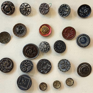 Pode incluir: Uma coleção de botões antigos em vários tamanhos e designs. Os botões apresentam padrões florais, geométricos e abstratos em tons de bronze, preto e dourado. Alguns botões têm um design em relevo, outros são planos.