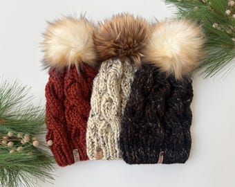 May include: Three chunky knit beanies featuring a cable knit pattern and large faux fur pom-poms. The hats are in deep red, speckled cream, and dark brown, each with a small rectangular leather-like tag attached to the bottom edge.