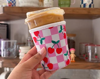 May include: A cylindrical neoprene iced coffee sleeve featuring a pink and white checkered pattern decorated with small red cherry illustrations. The sleeve is wrapped around a clear plastic cup filled with a light brown iced beverage.