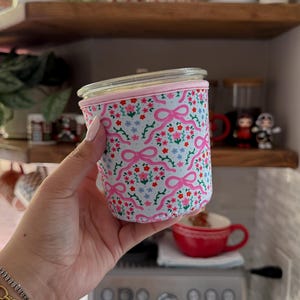 May include: A glass jar with a pink and floral patterned fabric sleeve. The sleeve features pink bows and small flowers on a light blue background. The jar is held in front of a kitchen setting with shelves and plants.