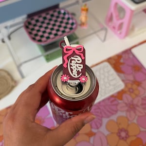 May include: A maroon soda can tab cover featuring a white straw, a pink bow, and two pink daisy flowers. The rectangular tag displays the white text Dr. Pepper in a script font. The accessory is attached to the silver pull tab of a red soda can.