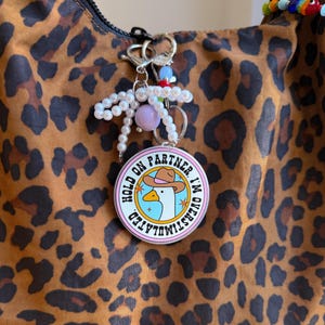 May include: A round keychain with the text "HOLD ON PARTNER, I'M OVERSTIMULATED" and a cartoon goose wearing a cowboy hat. The keychain is attached to a leopard print bag with a beaded charm.