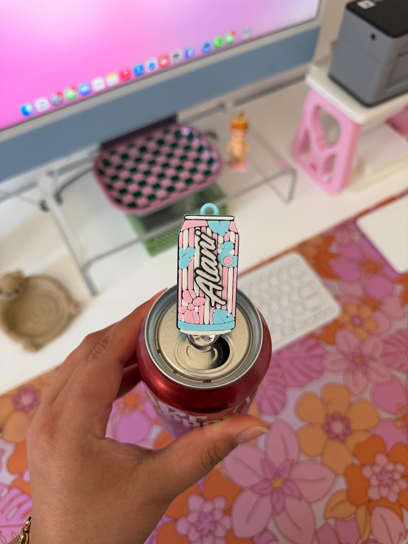 May include: A rectangular enamel pin shaped like a beverage can with a light blue pull tab at the top. The pin features a vertical striped pattern in pink and white, decorated with light blue and pink flower graphics and the text Alani in script.