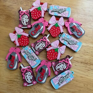 May include: A collection of colourful rubber charms featuring various designs including red strawberries with pink bows, soda cans labelled Dr. Pepper, Diet Coke, and Alani, all decorated with small pink bows and floral accents in various colours.
