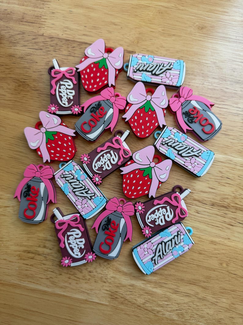 May include: A collection of colourful rubber charms featuring various designs including red strawberries with pink bows, soda cans labelled Dr. Pepper, Diet Coke, and Alani, all decorated with small pink bows and floral accents in various colours.