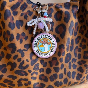 May include: A round keychain with the text "HOLD ON PARTNER I'M OVERSTIMULATED" and a cartoon goose wearing a cowboy hat. The keychain is attached to a bag with a leopard print pattern. The keychain also has a pearl and bead accent.