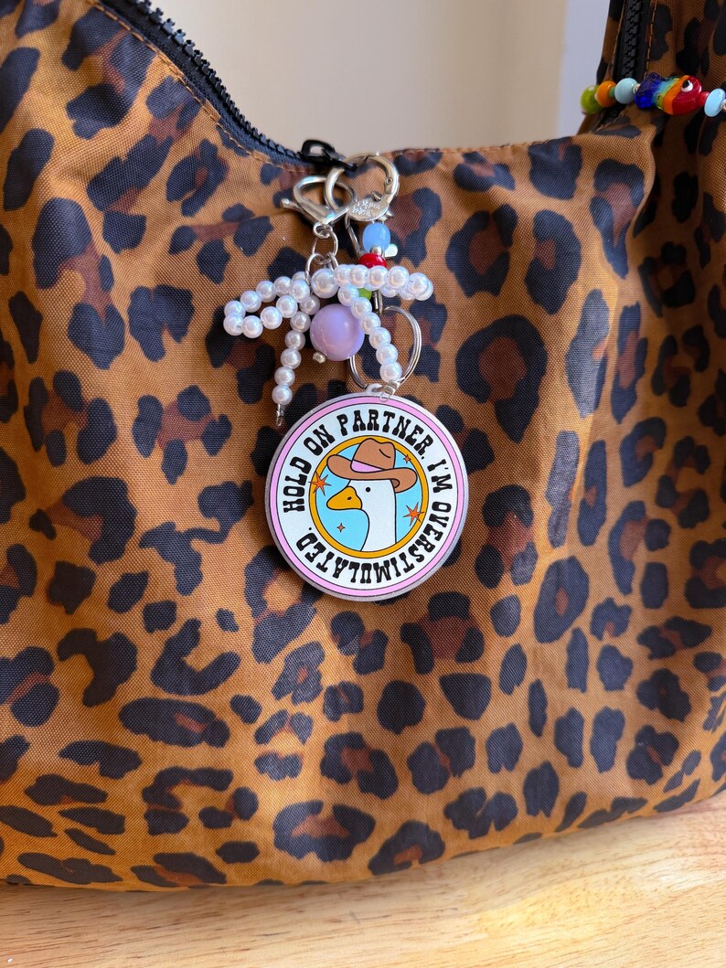 May include: A round keychain with the text "HOLD ON PARTNER I'M OVERSTIMULATED" and a cartoon goose wearing a cowboy hat. The keychain is attached to a bag with a leopard print pattern. The keychain also has a pearl and bead accent.