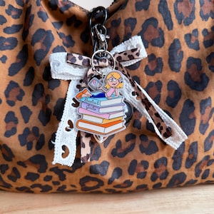 May include: A leopard print handbag featuring a decorative charm attached to a black metal clip. The charm displays a cartoon character lying on a stack of books with the text Bookish Girlie, accented by a white lace and leopard print ribbon bow.