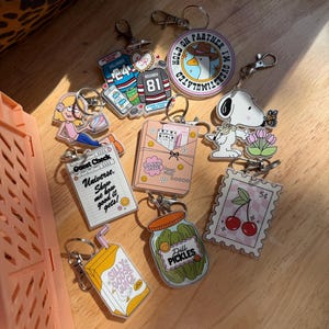 May include: A collection of acrylic keychains with various designs. Includes a Snoopy keychain, a jar of pickles, a juice box, a stamp with cherries, and keychains with text like "Guest Check" and "Silly Goose Juice".