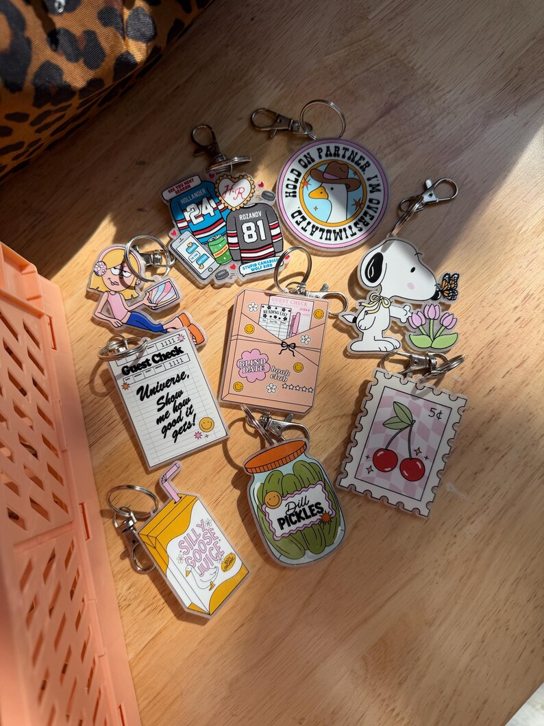 May include: A collection of acrylic keychains with various designs. Includes a Snoopy keychain, a jar of pickles, a juice box, a stamp with cherries, and keychains with text like "Guest Check" and "Silly Goose Juice".