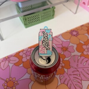 May include: A rectangular enamel pin featuring a stylised beverage can design with vertical pink and white stripes, light blue accents, and floral motifs. The word Alani is printed in a script font across the centre of the pin, which is attached to the tab of a metal soda can.
