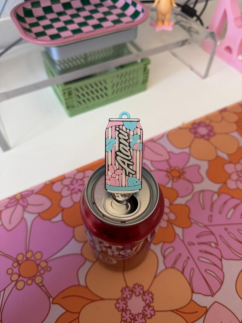 May include: A rectangular enamel pin featuring a stylised beverage can design with vertical pink and white stripes, light blue accents, and floral motifs. The word Alani is printed in a script font across the centre of the pin, which is attached to the tab of a metal soda can.