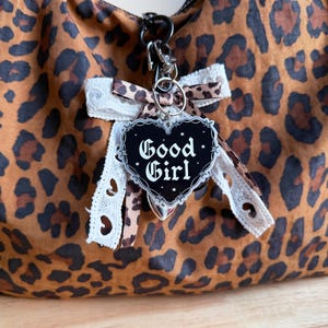 May include: A leopard print handbag featuring a black heart-shaped charm with the text Good Girl in white gothic lettering. The charm is accented with a barbed wire border and small stars, attached to a white lace bow and a leopard print ribbon.