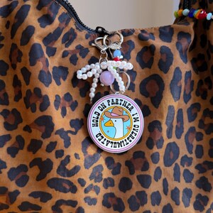 May include: A brown leopard print handbag with a silver zipper and a keychain. The keychain has a round charm with a cartoon goose wearing a cowboy hat and the text "HOLD ON PARTNER, I'M OVERSTIMULATED". The keychain also has pearls and colorful beads.