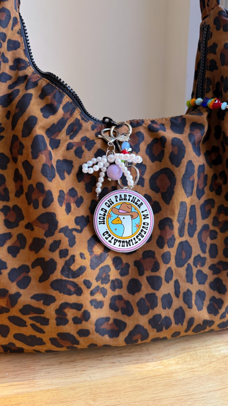 May include: A brown leopard print handbag with a silver zipper and a keychain. The keychain has a round charm with a cartoon goose wearing a cowboy hat and the text "HOLD ON PARTNER, I'M OVERSTIMULATED". The keychain also has pearls and colorful beads.