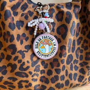 May include: A round keychain with the text "HOLD ON PARTNER I'M OVERSTIMULATED" and a cartoon goose wearing a cowboy hat. The keychain is attached to a beaded charm and a leopard print bag.