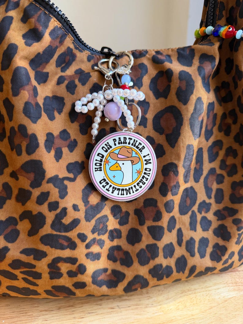 May include: A round keychain with the text "HOLD ON PARTNER I'M OVERSTIMULATED" and a cartoon goose wearing a cowboy hat. The keychain is attached to a beaded charm and a leopard print bag.
