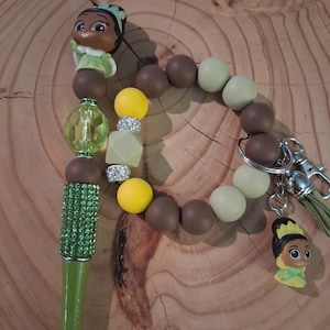 May include: A green pen with a jeweled barrel and a princess figurine on top. A matching beaded keychain with a princess charm and a wristlet are also shown. The beads are brown, yellow, and green.