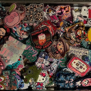 May include: A collection of colorful enamel pins with various designs, including cartoon characters, text, and patterns. The pins feature a variety of shapes and sizes, with text such as "CARE", "CHILD OF GOD", and "BURNT OUT".