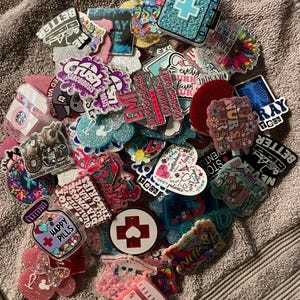May include: A collection of colorful medical-themed badge reels and charms. Designs include medical symbols, pills, and text like "Happy Pills", "Nurse Life", and "Crush Sarcoidosis". The items are in various shapes and sizes, with glitter and vibrant colors.