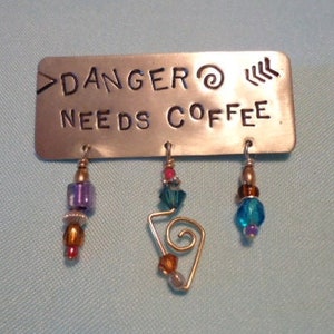 May include: A silver metal pin with the text "Danger Needs Coffee" and three dangling charms. The charms are a purple and brown bead, a blue and silver bead, and a blue and silver bead.