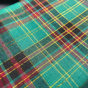 May include: Close-up of a green plaid fabric with black, red, and gold intersecting lines. The fabric has a woven texture and a diagonal pattern. The plaid design is a classic pattern.