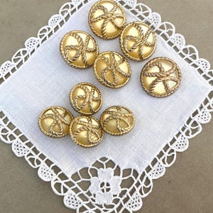 May include: Eight gold-colored buttons with a rope design around a pearl-like center. The buttons are arranged on a white, lace-edged cloth. The buttons are round and have a diameter of approximately 1 inch.