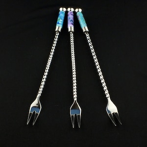 May include: Three cocktail forks with twisted silver stems and colorful, patterned handles. The handles feature blue, purple, and turquoise designs. The forks have a classic, elegant design, perfect for serving appetizers or garnishes.