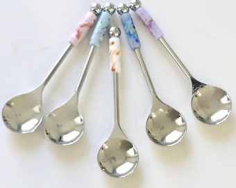 May include: A set of five silver-toned metal spoons arranged in a fan shape. Each spoon features a smooth, polished bowl and a decorative handle segment with a marbled pattern in pastel shades of pink, blue, white, and purple, topped with a small silver ball.